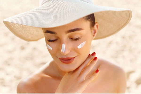 Zbliżenie na twarz młodej, uśmiechniętej kobiety, w kapeluszu na głowie i odrobiną niewchłoniętego jeszcze kremu z filtrem na policzkach